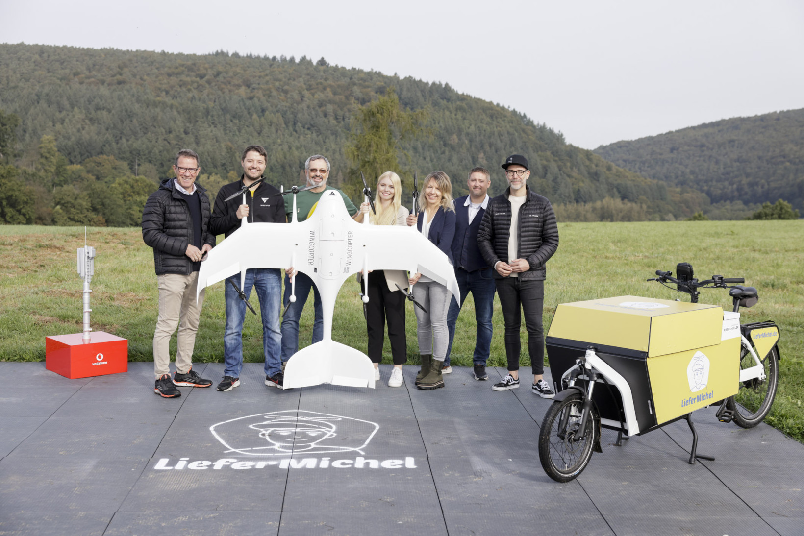 A group of people standing behind a Wingcopter drone with cargo bike to the side